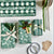 Flat-lay of green wrapping paper and ribbon supplies with three wrapped gift boxes, tape, kraft tags, and a pencil, arranged on a light neutral background.