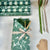 A wrapped gift box with a green floral paper pattern, tied with a beige polka-dot ribbon and decorated with pine branches and wheat, shown on a white surface with rolls of matching wrapping paper and ribbon.