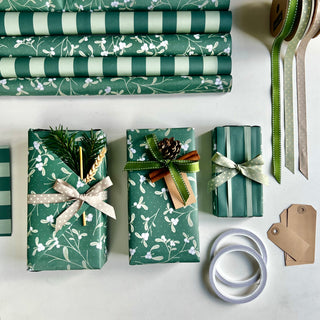 A styled flat-lay of wrapping paper and gift wrap supplies on a light surface. Several wrapped gift boxes are shown with green floral and striped wrapping paper, tied with ribbons and decorated with pine branches, a gold stick, and a pinecone.