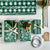 A styled flat-lay of wrapping paper and gift wrap supplies on a light surface. Several wrapped gift boxes are shown with green floral and striped wrapping paper, tied with ribbons and decorated with pine branches, a gold stick, and a pinecone.
