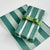 Green and white striped wrapping paper with a floral pattern, shown wrapped around a gift box tied with a green ribbon, with additional rolls of the same paper in the background.