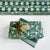 Green floral wrapping paper with white leaf motifs, shown wrapped around a gift box tied with a green ribbon and cinnamon sticks, with additional rolls of the same pattern in the background.