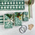 Flat-lay of green wrapping paper and ribbon supplies with three wrapped gift boxes, tape, kraft tags, and a pencil, arranged on a light neutral background.