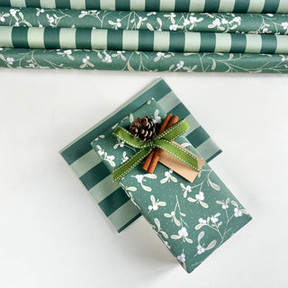 Green and white holiday wrapping paper shown both rolled in the background and wrapped around a gift box with a green ribbon, cinnamon sticks, and a pinecone on a white surface.