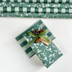 Green and white holiday wrapping paper shown both rolled in the background and wrapped around a gift box with a green ribbon, cinnamon sticks, and a pinecone on a white surface.
