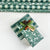Green and white holiday wrapping paper shown both rolled in the background and wrapped around a gift box with a green ribbon, cinnamon sticks, and a pinecone on a white surface.