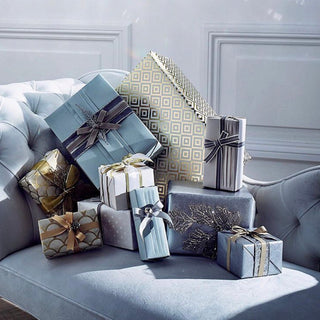A collection of wrapped Christmas gift boxes in shades of blue, gray, and gold, styled on a tufted light blue sofa against a gray paneled wall.