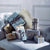 A collection of wrapped Christmas gift boxes in shades of blue, gray, and gold, styled on a tufted light blue sofa against a gray paneled wall.