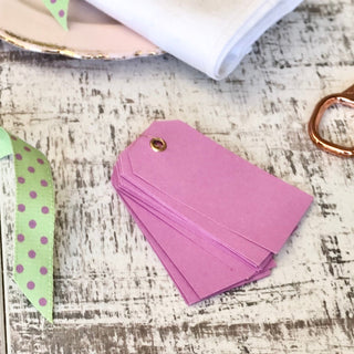 A stack of pink paper gift tags with a gold grommet, arranged on a distressed white wooden surface with decorative props including a green polka-dot ribbon, a folded white cloth, and a rose gold ring.